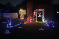A nighttime photo of a Nightmare Before Christmas–themed front yard display featuring a cemetery scene with glowing tombstones, string lights outlining paths in the grass, a tall Christmas tree decorated with pumpkins and lights, Jack Skellington dressed as Santa near a doghouse, and Zero the ghost dog floating under a lit stone archway. Patches of artificial snow cover parts of the yard.
