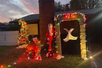 A close-up view of a Nightmare Before Christmas–themed yard display taken at dusk. Jack Skellington dressed as Santa stands beside Zero’s doghouse, both wrapped in colorful Christmas lights. Zero the ghost dog floats under a stone archway decorated with multicolored lights and patches of snow. To the left, a tall Christmas tree is trimmed with glowing lights and pumpkin ornaments, with artificial snow resting on its branches.