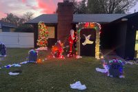 A daylight photo of a Nightmare Before Christmas–themed front yard display. Jack Skellington in his Santa outfit stands beside Zero’s decorated doghouse, surrounded by colorful Christmas lights and patches of artificial snow. Zero the ghost dog floats under a stone archway wrapped in multicolored lights. A tall Christmas tree decorated with glowing lights and pumpkin ornaments stands to the left, and several light-lined tombstones are arranged across the lawn.